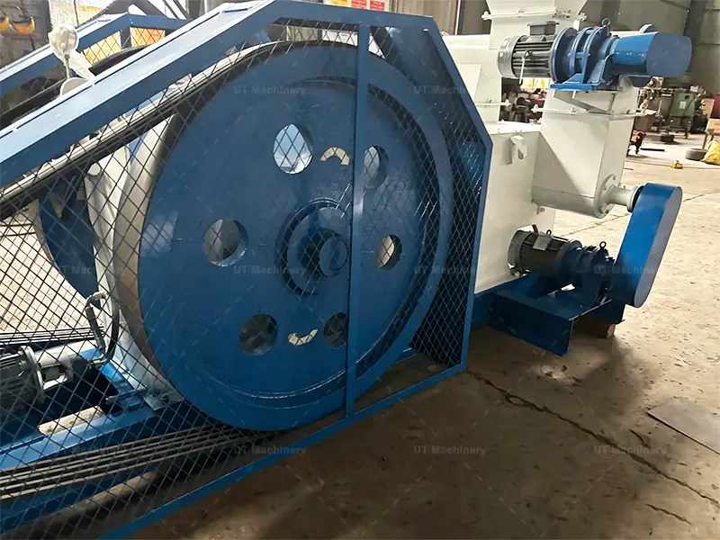 Gran volante de inercia azul y sistema de correa de transmisión con una malla de protección de seguridad en una máquina briqueteadora de biomasa industrial.