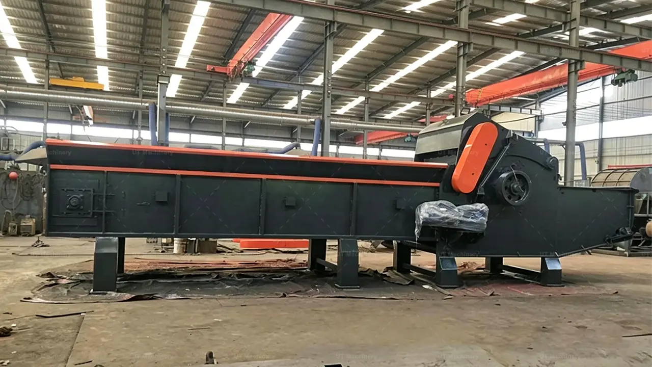 Vista de perfil lateral a larga distancia y a la altura de los ojos de una enorme astilladora de madera industrial gris oscuro dentro de un taller bien iluminado. La máquina cuenta con una tolva de alimentación baja y muy larga a la izquierda con un sistema de arrastre de tablillas y cadenas, que conduce a la gran unidad de procesamiento a la derecha.